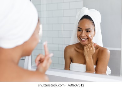 Portrait Of A Young Smiling Woman Applying Moisturiser To Her Face In The Bathroom