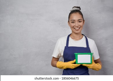 Portrait Of Young Smiling Woman, Cleaning Lady In Uniform And Yellow Rubber Gloves Holding Green Plastic Box With Washing Capsules While Standing Against Grey Wall