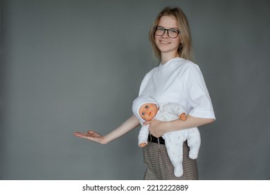 Portrait Of Young Smiling Woman Holding Baby Doll In White Clothes, Stretching Hand Aside With Open Palm On Grey Background. Motherhood, Childbirth, Breastfeeding, Breast Model, Fake, Education. 