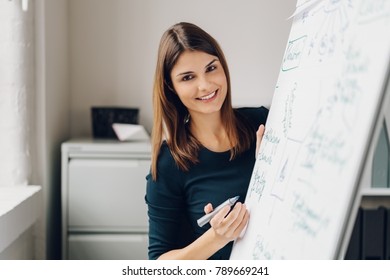 Portrait Of Young Smiling Woman Holding Marker Pen While Standing By Flip Chart In Office