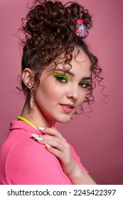 Portrait of young smiling woman on pink background. Female with unusual green eyes shadows makeup, curly hair and earrings. Hands are near face. - Shutterstock ID 2245422397