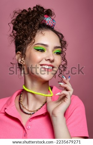 Portrait of young smiling woman on pink background. Female with unusual green eyes shadows makeup, curly hair and earrings. Hands are near face.