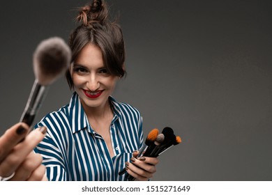 Portrait Of Young Smiling Woman In Striped Blouse With Makeup Brushes In Her Hands And Showing One Defocused Brush In Camera Isolated