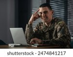 Portrait of a young soldier in a military uniform working on a laptop while sitting at a table at home.