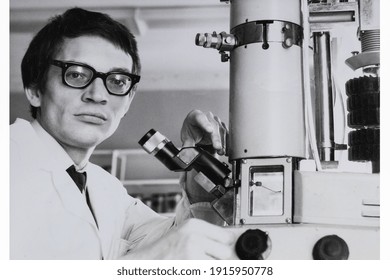 Portrait Of Young Soviet Guy With Glasses And White Medical Gown, With Microscope. Vintage Black And White Paper Photo. Early 1980s. Old Surface, Soft Focus. Transferred Property, Family Archive.