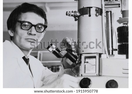 Portrait of young Soviet guy with glasses and white medical gown, with microscope. Vintage black and white paper photo. Early 1980s. Old surface, soft focus. Transferred property, family archive.