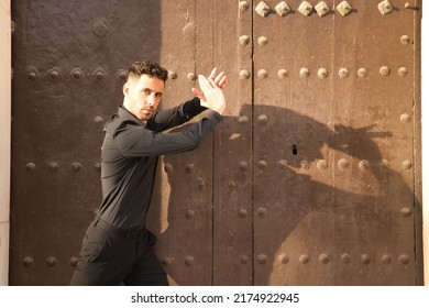 Portrait Of Young Spanish Man, Wearing Black Shirt And Pants, Dancing Flamenco In The Street Next To A Large Wooden Door Casting His Shadow On It. Typical Spanish Concept, Art, Dance, Culture.