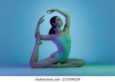 Portrait of young sportive girl doing stretching exercises isolated over blue studio background in neon light. Developing flexibility. Concept of sport, strength, health, hobby, youth, lifestyle, ad