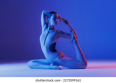 Portrait of young sportive girl doing legs stretching exercises isolated over blue studio background in neon light. Concept of sport, health, hobby, youth, strength, lifestyle, ad