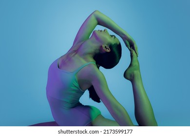 Portrait of young sportive girl doing stretching exercises isolated over blue studio background in neon light. Flexible body. Concept of sport, health, hobby, youth, strength, lifestyle, ad