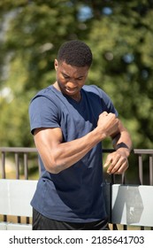 Portrait Of Young Sportive Strong Tall Handsome African American Black Skin Man With Tense Muscles Looks At His Hand, Green Tree, Building Are On Background. Vertical Plane. High Quality Photo.