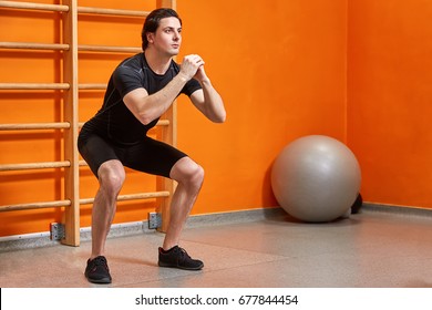 Portrait Of Young Sportsman In The Black Sportwear While Making Squat Against Bright Orange Wall In Gym.