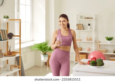 Portrait of a young sporty slim woman wearing sportswear standing at home after working out with glass of water and vegetables on the table. Healthy food eating, nutrition and lifestyle concept. - Powered by Shutterstock