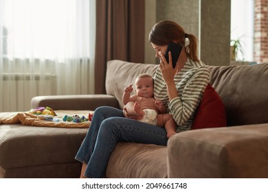 Portrait Of Young Stressed Mother Sitting Holding Baby In Arms, Calling To Clinic To Set Appointment To Family Doctor. Desperate Woman Trying To Understand What Is Wrong With Her Child.