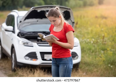 Portrait Of Young Stressed Woman Standing At Broken Car And Reading Owner Manual