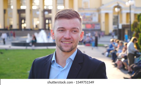 Portrait Of Young Stylish Businessman Looking And Smiling Into Camera With Positive Emotions. Confident Handsome Man Straightening Hair And Looking Around. Face Of Smiling Guy Posing Outdoor. Slow Mo