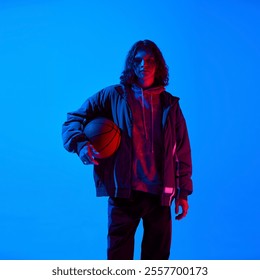 Portrait of young stylish man with long hair, holding basketball, wearing hoodie and jacket against blue studio background in neon light. Concept of sport, youth, street style, modern fashion - Powered by Shutterstock