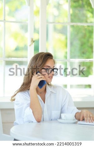                             Portrait of a young successful businesswoman sitting at a table in a home white office, working on a laptop, talking on a mobile phone. Smiling, cute girl in glasses wearin