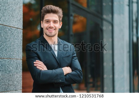 Portrait of young successful confident businessman in the city on office building background. Man in business suit looking to camera and smiling. Portraiture of handsome guy