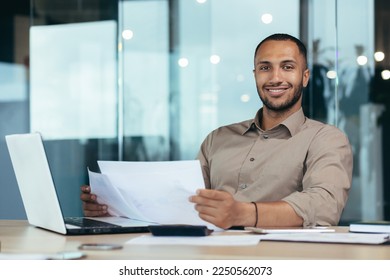 Portrait of young successful hispanic businessman inside office, man smiling and looking at camera, paper worker happy with achievement results sitting at workplace with laptop. - Shutterstock ID 2250562073