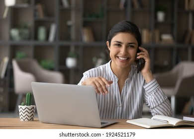 Portrait of young successful Indian businesswoman sit at desk at home office work on laptop talk on cellphone. Smiling millennial ethnic woman use computer have smartphone call with client.