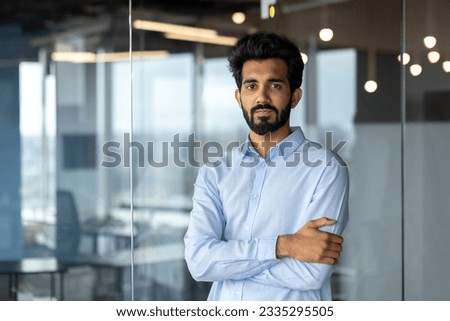 Portrait of a young successful Indian male programmer, developer standing in an office center, crossing his arms over his chest and looking seriously at the camera.