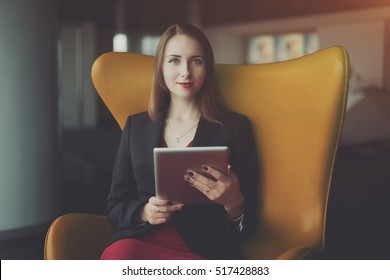 Portrait Of Young Successful Woman Entrepreneur In A Red Dress And Jacket Sitting On Orange Armchair And Working On Her Digital Tablet, Office Interior