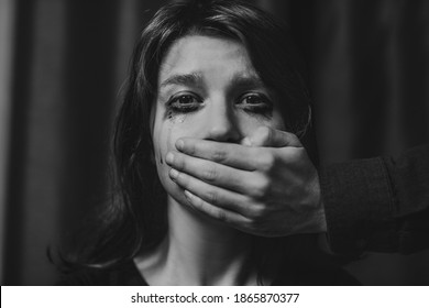 Portrait of a young suffering sad woman with tears in her eyes, a man closes her mouth with his hand. Domestic violence, crying, religion, disagreement, fight, divorce, beating a weaker person, dark.