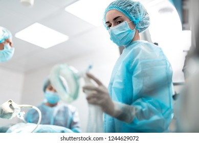 Portrait Of Young Surgical Nurse In Sterile Glove Holding Respiratory Equipment For Patient. Focus On Lady Face In Protective Mask