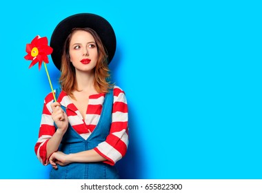 Portrait Of Young Surprised Red-haired White European Woman In Hat And Red Striped Shirt With Jeans Dress With Pinwheel On Blue Background