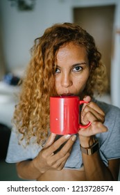 Portrait Of A Young, Tanned And Sexy Indonesian Asian Woman Enjoying A Hot Mug Of Tea On Her Own At Home On A Morning. She Has Dyed Frizzy Hair And Is Wearing Comfortable Home Clothes. 