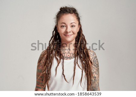 Portrait of young tattooed caucasian woman with dreadlocks wearing white shirt, smiling at camera while posing isolated over grey background. Front view. Horizontal shot