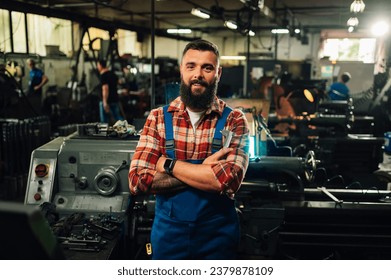 A portrait of a young tattooed machinist with a beard posing in a factory in front of a lathe machine, crossed arms. He is wearing a shirt and overall pants.  - Powered by Shutterstock