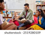 Portrait of young teacher reading book to primary students in school library. Man reading a fairy tale to a group of multiethnic schollboys and schoolgirls. Librarian at primary school reading a story