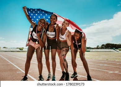 Portrait Of Young Team Of Female Athletes Enjoying Victory. Diverse Group Of Runners With Medals Celebrating Success Holding A USA Flag On Racetrack.