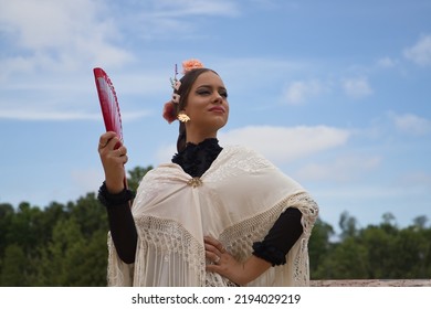 Portrait of young teenage girl in black dance dress, white shawl and pink carnations in her hair, dancing flamenco with a red fan. Concept of flamenco, dance, art, typical Spanish dance, fan.