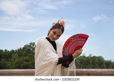 Portrait Of Young Teenage Girl In Black Dance Dress, White Shawl And Pink Carnations In Her Hair, Dancing Flamenco With A Red Fan. Concept Of Flamenco, Dance, Art, Typical Spanish Dance, Fan.