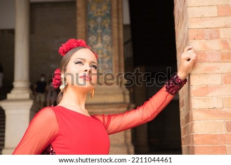 Portrait of young teenage woman in red dance suit with red carnations in her hair doing flamenco poses leaning against a brick wall. Flamenco concept, dance, art, typical Spanish dance.