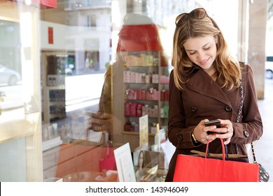 Portrait of a young teenager tourist visiting the city and carrying paper shopping bags while leaning on a fashion store window, using her smartphone device and smiling.