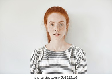 Portrait of young tender redhead teenage girl with healthy freckled skin wearing striped top looking at camera with serious or pensive expression. Caucasian woman model with ginger hair posing indoors