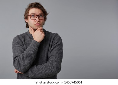Portrait of young thinking man with glasses wears in gray sweater, stands over gray background with copy space on the right side, touches chin and looks away. - Shutterstock ID 1453945061