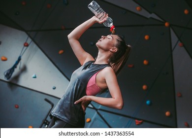 Portrait Of Young Tired Sweaty Fitness Girl Drinking Water From Bottle While Standing In Dark Gym. Beautiful Wet Sports Woman Pouring Water On Her Body After Hard Workout Training. Sexy Athlete Girl