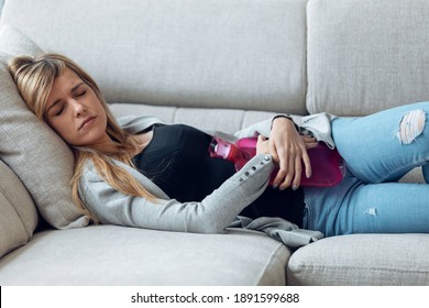 Portrait Of Young Tired Woman Lying On Sofa With Hot Water Bottle For Stomachache In Living Room At Home.