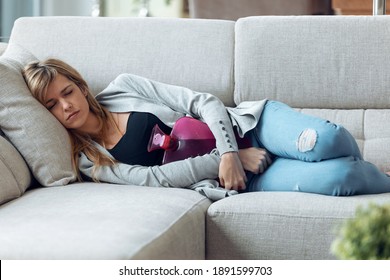 Portrait Of Young Tired Woman Lying On Sofa With Hot Water Bottle For Stomachache In Living Room At Home.