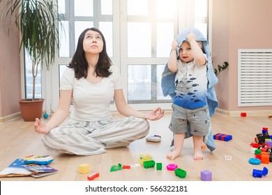 Portrait Of Young Tired Yoga Mom Spending Time With Her Little Baby Boy, Relaxing While Her Kid Son Playing Games, Throw Toys And Arrange A Mess At Home