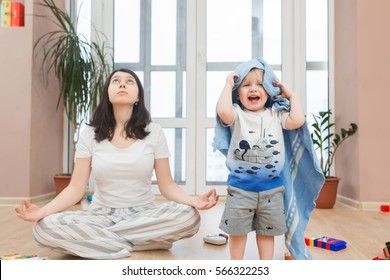 Portrait Of Young Tired Yoga Mom Spending Time With Her Little Baby Boy, Relaxing While Her Kid Son Playing Games, Throw Toys And Arrange A Mess At Home