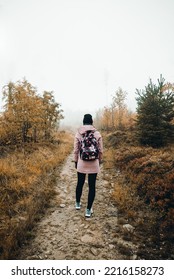 Portrait Of Young Tourist Woman With Backpack In Colorful Autumn Misty And Foggy Forest. Female Turist During The Hike - Vertical Photo From Back.