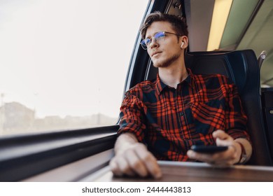 Portrait of a young traveler sitting in train near window, looking trough it and listening to music. A young hipster guy is traveling by railroad transportation and looking through window. Copy space. - Powered by Shutterstock