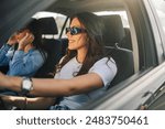 Portrait of young trendy attractive female driver driving a car with hands on steering wheel and smiling while her friend is sitting next to her and enjoying a ride. Two friends in car on a road trip.