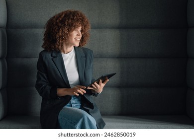 Portrait of young trendy businesswoman with curly hair sitting at modern workspace with tablet in hands and smiling.Happy fashionable millennial businesswoman posing at minimalistic office with tablet - Powered by Shutterstock